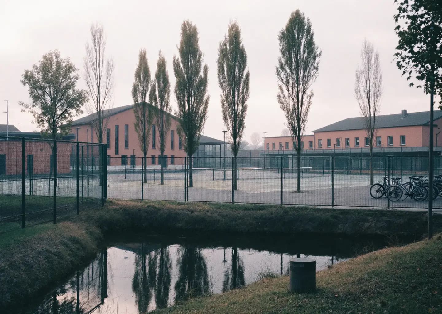 Parking d'un complexe sportif municipal, courts grillagés et peupliers au bord d'une rivière